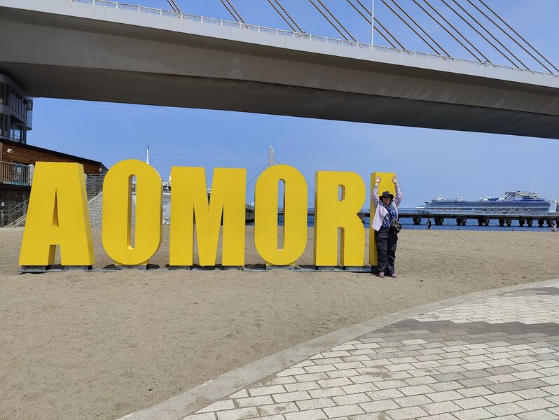 Bright yellow letters spelling AOMORI, with Holly standing in front of the I with her arms raised to mimick it.