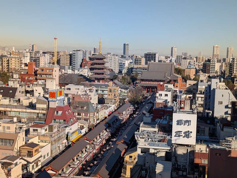 High up view of Buddhist temple Sensoji, a 5 storey pagoda, and the shopping street leading up to it. The temple, pagoda and vendor stalls all have the same tile roof with vermilion red trim. The lighting and angle make it look like it's in miniature.
