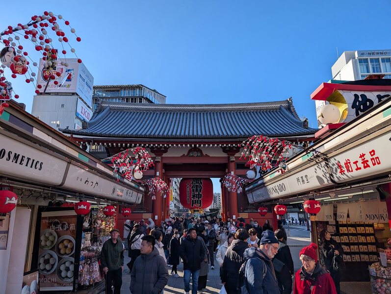 Entrance gate and some shops in the shopping street leading up to Sensoji. The gate has a tile roof, vermilion supports, and a giant red lantern in the middle. The shops have an open front with food and souvenirs for sale. Mom and Dad are visible in the foreground.