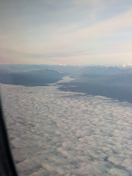 A bunch of clouds seen from above, clearing to reveal mountains, an inlet, and high mountains beyond