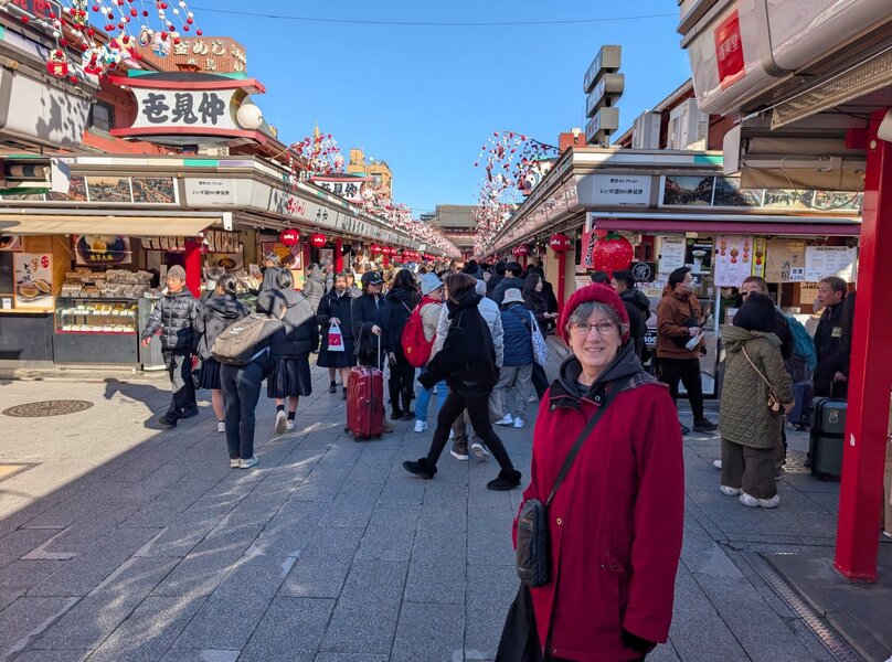 Halfway down the shopping street, with the temple in the background. Mom is posed nicely in a red coat and red toque in front of some schoolchildren and stalls with food for sales (tiny pancakes, and chocolate covered fruit on a stick)