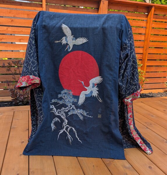 Indigo happi style coat with a large pattern of two cranes flying in front of a red sun above some Japanese woodblock-style trees. The sleeves have a different pattern of small white stripes, and the cuffs and neckline have a fancy red fabric with an embroidered pattern.