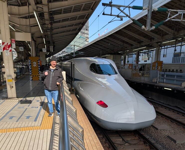 Andrew standing on a train platform next to the front of a shinkansen train. Andrew looks excited, and the train looks very sleek and modern. Power lines run above and the train station has an industrial vibe.