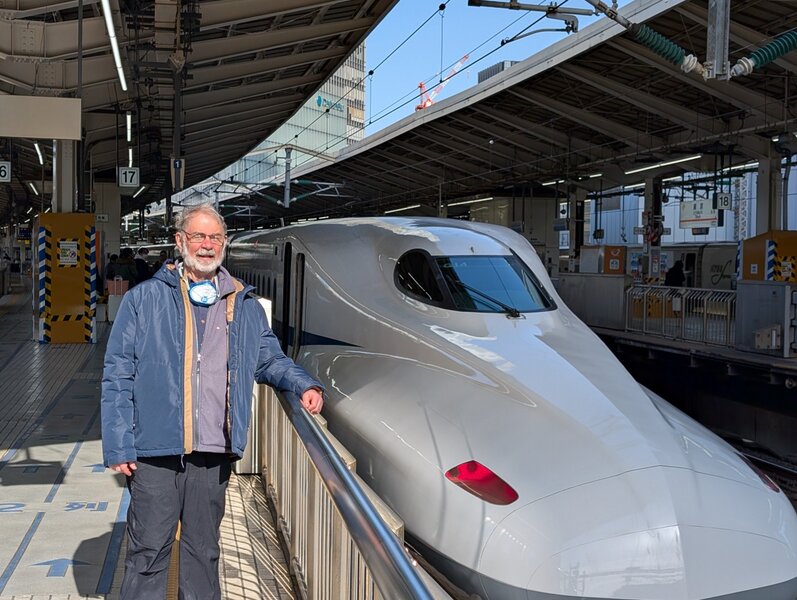 Dad standing on the same train platform next to the front of a shinkansen train. He's squinting into the sun next to the sleek and modern train.